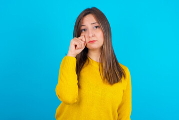 Disappointed dejected Young arab woman wearing knitted sweater over blue backgtound wipes tears stands stressed with gloomy expression. Negative emotion