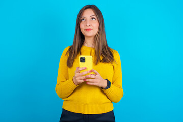 Young arab woman wearing knitted sweater over blue backgtound holds mobile phone uses high speed internet and social networks has online communication. Modern technologies concept