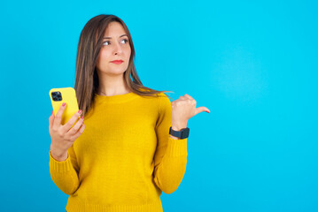 Young arab woman wearing knitted sweater over blue backgtound points thumb away and shows blank space aside, holds mobile phone for sending text messages.