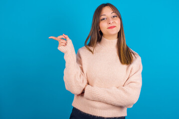 Fototapeta premium Young arab woman wearing knitted sweater over blue backgtound points to side on blank space demonstrates advertisement. People and promotion concept