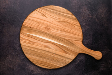 Round empty wooden charcuterie serving board with handle on old dark brown shabby table. View from above. Top view. Food background. Template with copy space for text. Flat lay, mockup. Layout, frame.
