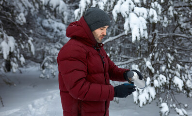 Fototapeta na wymiar Handsome young Caucasian man with bearded chin in grey sport hat and red down jacket standing in winter snowy forest pouring some hot drink into cup from thermos to warm himself up during his trip