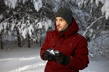 Fototapeta na wymiar Handsome man in winter clothes holding vintage camera in hands, looking for best view to take picture, enjoying winter landscape of nature, snowy trees, pines, spruces, breathing fresh frosty air