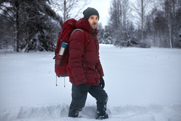 Horizontal outdoor portrait of male hiker in warm clothes and big red backpack with thermos in its pocket laying path through snowdrifts, walking alone, enjoying fresh air, beautiful landscape