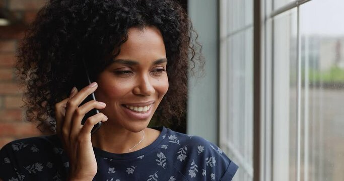 Positive smiling Afro American woman stand by light window hold mobile phone at ear make answer call speak to friend husband listen to good news. Friendly young lady chat talk on cell discuss affairs