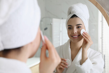 Beautiful young woman cleaning her face with cotton disk near mirror in bathroom