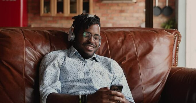 Smiling African man in wireless earphones relax on sofa using phone audio app watch movie video clip select music track in online playlist. Happy male hipster spend free time playing web game on cell