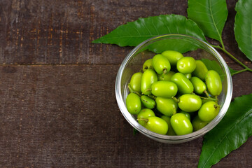 Fresh Neem fruit on transparent glass bowl and neem green leaf on rustic wooden background.
