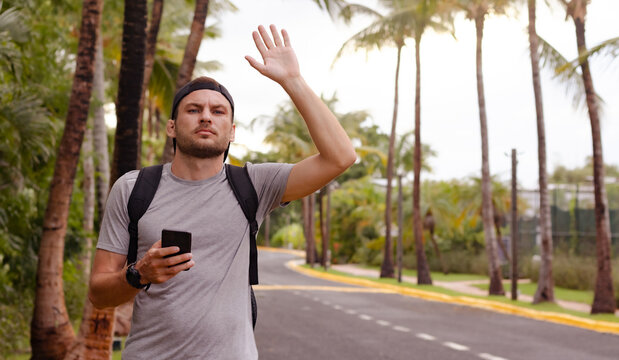 Man traveler stands by the road and waving his hand, hold smartphone in another hand. Man hailing a taxi in Dominican Republic.