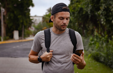 Man backpacker in cap stands on the road and looks at the screen of mobile phone in his hand. Hiking, navigation, tourism, travel concept.