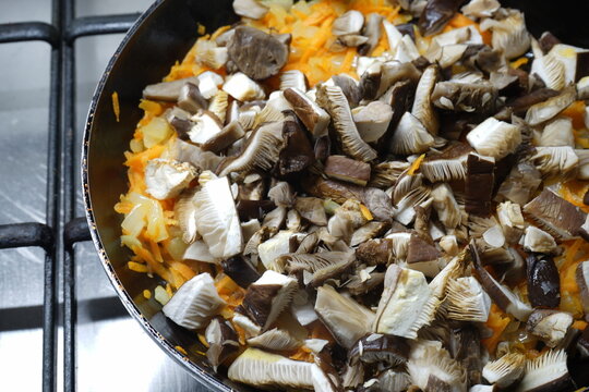 Oyster mushrooms are cut into strips, onions and chopped carrots are fried in a frying pan. There is a cast-iron frying pan with mushrooms, onions and carrots on the gas stove.