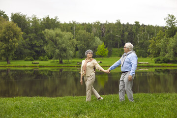 Naklejka na ściany i meble Senior man and woman holding hands in the park, walking outdoors, happy elderly family. Elderly people walk near the river. Green grass, summer.