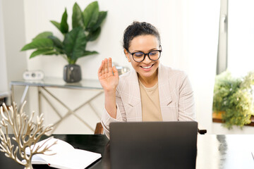 Video meeting concept. Cheerful middle-aged woman holding video call on the laptop at office, waving hi into webcam, greeting online interlocutors, involved online video meeting with work team © Face Stock