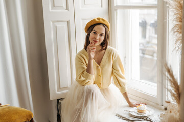 Young lovely fair-skinned brunette woman sits on windowsill in afternoon looking into camera. Model girl in yellow beret, jacket and skirt starts her morning with pleasant views from window.