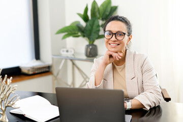 Confident middle-aged woman work with a laptop at the modern office, female employee in smart casual wear looks at the camera and smiles, typing messages, answering email, front view