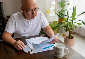A senior man sitting by a table calculating the raising cost of energy and tax bills. Inflation and...