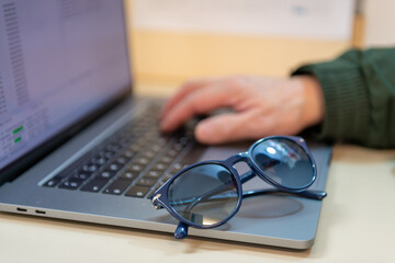 A person using a laptop with a hand on the key board and a pair of sunglasses. Online scams, cyber attacks, fraud concept.