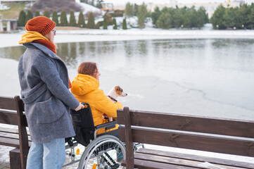 Caucasian woman in a wheelchair and her friend are sitting by the lake with ducks in winter.