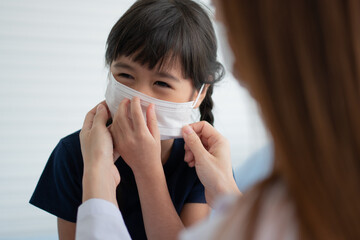 Asian parents wear face masks to little daughter for protection against coronavirus covid 19 before going to school. Mom teaches daughter to put on a medical mask