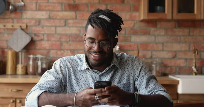Smiling African American male hipster in eyewear use phone app in kitchen play mobile game write message in chat buy goods at web shop. Happy young man user browse social media dating online via cell
