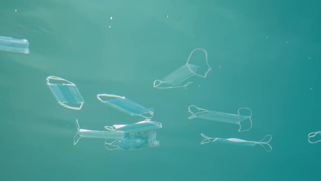 Used medical masks drifts underwater in blue water column. Coronavirus is contributing to pollution, as discarded masks clutter polluting seas and ocean along with plastic trash. Close up, slow motion