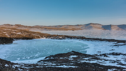 Two frozen lakes are located nearby and are separated by a strip of land. Snow patterns on ice. A mountain range against a blue sky. Baikal.