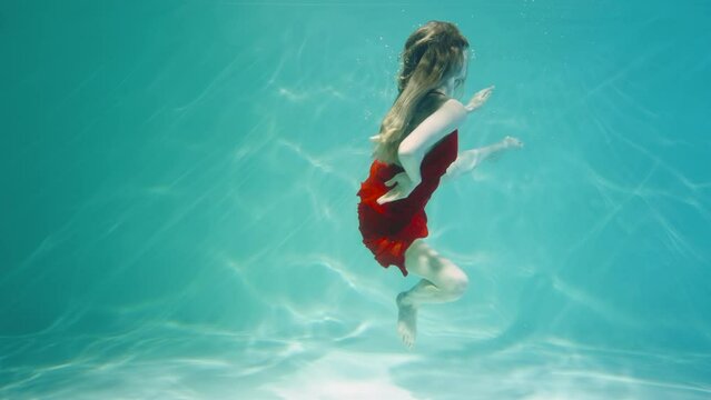 Young woman in red chiffon costume dancing underwater in blue water column with glare. Female dancer dancing and floating in sea water, floats to surface for breath of air. Close up in slow motion.