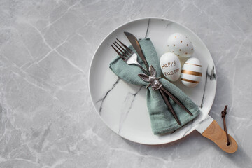 Banner. Table setting. A fashionable marble plate with a rabbit on a napkin, Easter eggs and feathers on a gray background. Top view. Happy Easter holiday concept for cafes and restaurants.