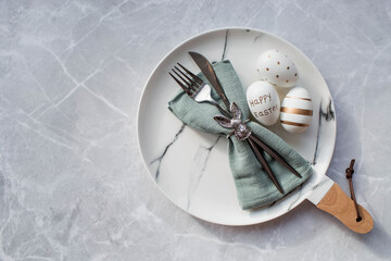 Banner. Table setting. A fashionable marble plate with a rabbit on a napkin, Easter eggs and feathers on a gray background. Top view. Happy Easter holiday concept for cafes and restaurants.