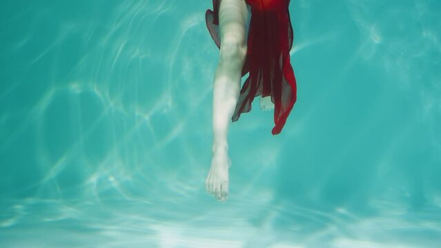 Underwater shot of woman dancing in red chiffon costume. Female dancer gracefully moves legs in blue water column with glare. Woman subaquatic shot, close up in slow motion.