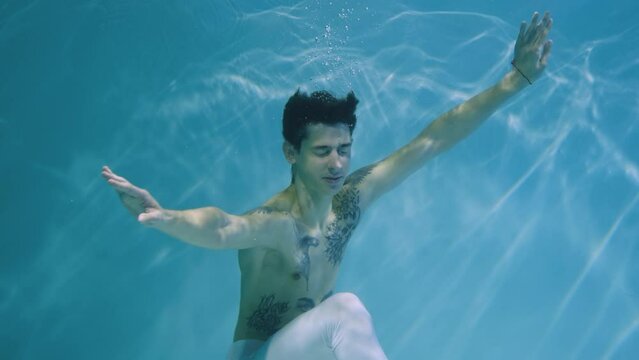 Underwater shot of man with a halfnaked torso gracefully dancing modern ballet choreography. Male dancer moves in blue water column with glare. Subaquatic shot in pool, close up in slow motion.
