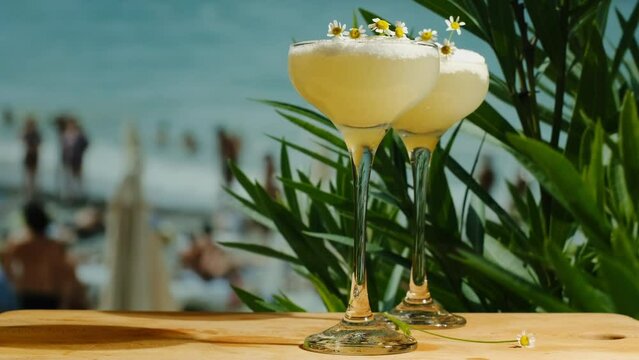 Beach bar. Foamy alcoholic lemon cocktails on table bar, sea on background. Two drinking glasses of pisco sour cocktails decorated fresh chamomiles. Summer sea resort concept.