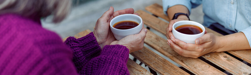 Mature women best friends with cups of fresh coffee sit at wooden table on outdoors cafe terrace on...