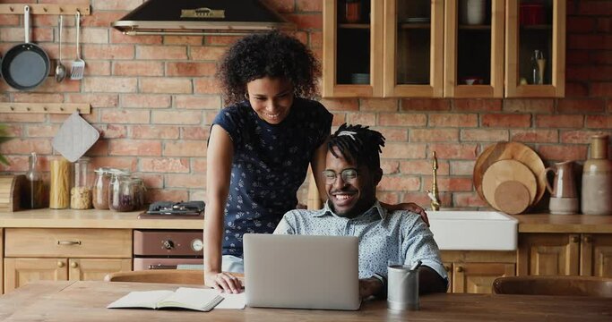 Young Black husband sit by pc at kitchen talk to wife standing behind listen to advice purchase proposition choosing item at ecommerce shop. Friendly couple discuss plans internet offers buying goods
