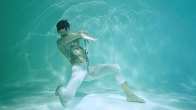 Underwater shot of man with a halfnaked torso wrapped in chains dancing modern ballet choreography. Male dancer moves in blue water column with glare. Subaquatic shot in pool, close up in slow motion