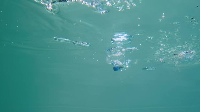 Discarded plastic bottles and cups drifting underwater in blue water column with glare. Plastic garbage environmental pollution problem in seas and ocean. Ecological catastrophy. Close up, slow motion