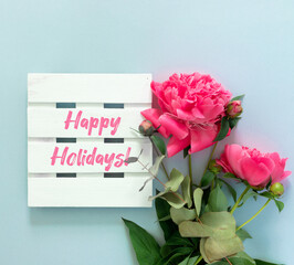 Bouquet of beautiful pink peonies with gift boxes in paper wrapping. White wooden board with text Happy holiday!