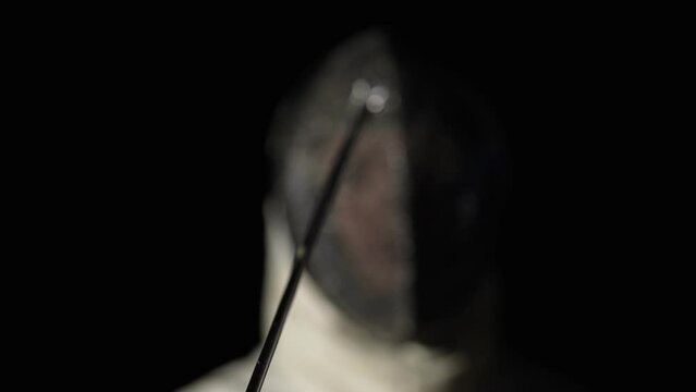 Portrait of young woman fencer in protective helmet pointing rapier at camera. Sportswoman in white uniform poses in dark studio on black background with backlight. Slow motion. Close up.