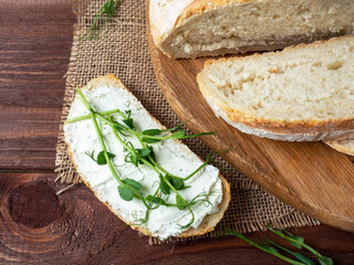 Close-up of a still life made of fresh crusty bread. A slice of bread smeared with cottage cheese and decorated with micro greenery. Linen napkin brown wooden background, top view, flat lay