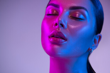 Glossy, wet skin makeup. Fashion model woman face in bright neon colourful lights, beautiful woman, UV, ultraviolet trendy make-up. Art design, professional make up. Glittering, shine make-up