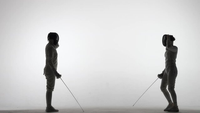 Silhouettes of two female swordsmen fighting a duel. Athletes cross rapiers and deliver attacking blows. The girls pose in a dark studio on a white background in a spotlight spot. Slow motion.