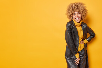 Fashionable positive woman dressed in jumper leather coat and trousers looks gladfully away has...