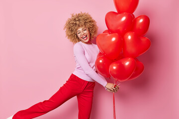 Positive carefree woman dances with bunch of heart balloons enjoys Valentines celebration has fun on party dressed in neat clothes laughs joyfully isolated over pink background. Holidays concept