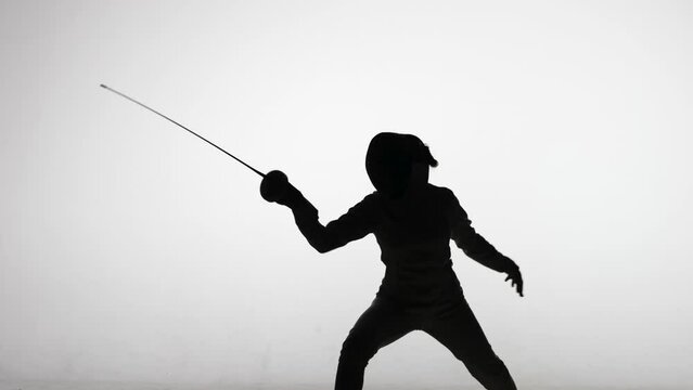 Silhouette of young woman swordsman throwing attacking blows with sword. Sports training in fencing school. Girl athlete posing in dark studio on a white background in spot of spotlight. Slow motion.