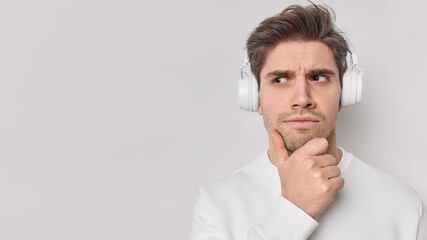 Fototapeta premium Serious male student listens educational content holds chin wears wireless headphones with good sound quality on ears looks away with attentive expression isolated over white wall copy space