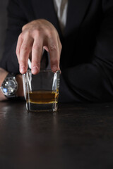 Hand of a man in a white shirt and dark suit holds a glass of whiskey on the bar