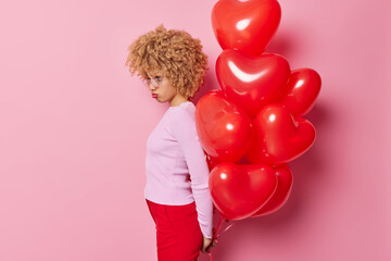 Offended young woman with curly hair stands sideways at camera holds bunch of inflated balloons behind back feels stressed and lonely isolated over pink background. Sad Valentines Day concept