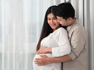 Fototapeta premium Portrait of Asian couple hugging and both putting hands on tummy of pregnant woman.