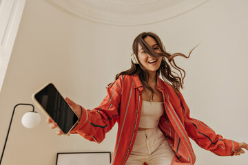 Energetic young model with wireless headphones is having lot of fun in closed white room. Dark-haired girl wear casual clothes holds phone in her hand and smiles cutely at camera. - obrazy, fototapety, plakaty