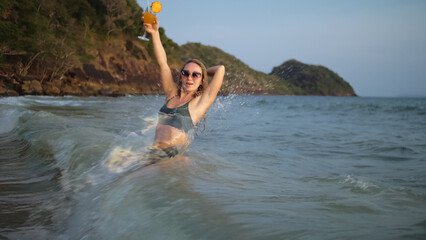 Sexy hot woman stand knee-deep in sea on golden sunset. Girl on tropical beach in green swimsuit having fun and waving his hands, drinks her orange cocktail Pina Colada. Big waves hitting buttocks